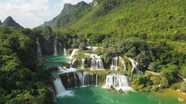 Stunning aerial footage of the Ban Gioc Waterfall cascading over multiple tiers surrounded by lush green forest and limestone mountains in Cao Bang province, northern Vietnam.