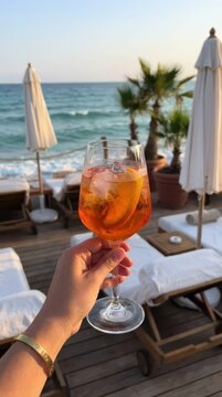 Hand lifting glass with orange cocktail at seaside lounge for summer cocktail banner and social media background with sunset view