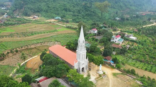 Drone footage showing a Catholic church with a tall white steeple and red roof, surrounded by lush green fields, scattered houses, and forested hills in the rural village of Bach Xa, Vietnam.