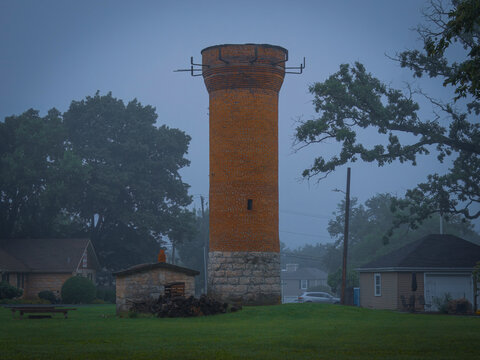 An old vintage red brick water tower stands tall in a misty rural residential neighborhood surrounded by lush green trees