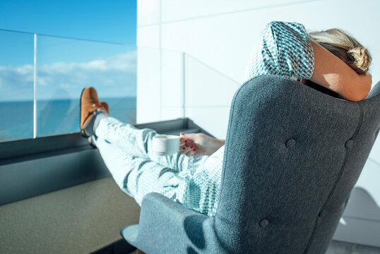 Woman relaxing in a gray armchair on a balcony overlooking the ocean, holding a white cup, wearing a patterned outfit with a scenic view of blue sky and water