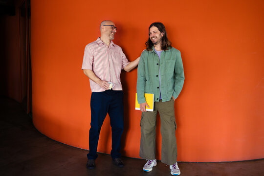 Two Friends Laughing During Casual Conversation Against Orange Wall