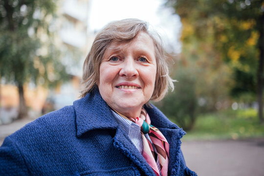 Elderly woman with short gray hair wearing a blue coat and patterned scarf smiles while standing outdoors in a park with trees and buildings in the background