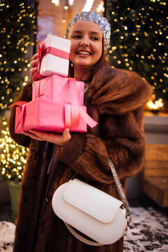 Young woman with long brown hair wearing a fur coat holds colorful wrapped gifts while standing outdoors in a festive setting with lights and decorations