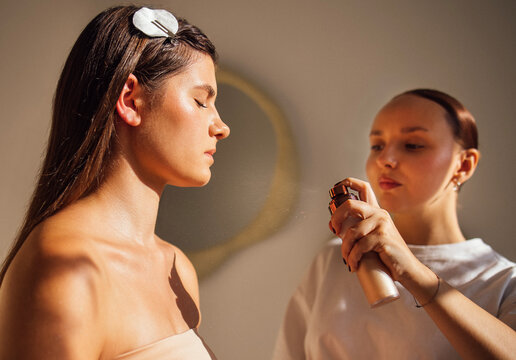 Young woman receiving beauty treatment from a professional makeup artist, showcasing skincare application in a bright, modern salon environment with soft lighting and elegant decor