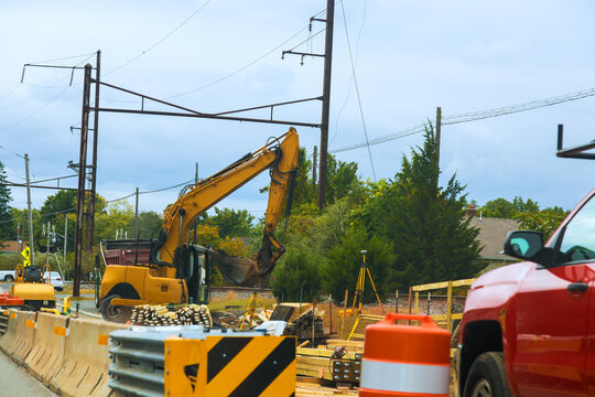 Digger is working on side of road while cars pass by there are construction materials barriers nearby.