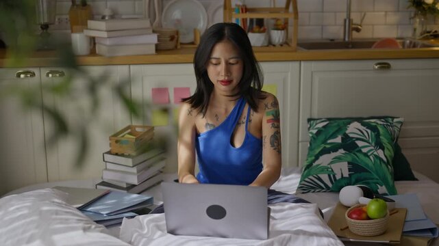 Freelancer woman Gen Z sitting on bed and typing on a laptop in apartment.asian female remote work in kitchen at home.digital nomad,online work,remote working and video conference training concept