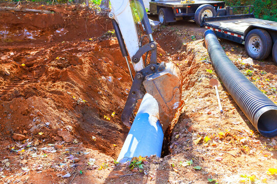 Excavator machine is digging into ground to install sewer pipe in construction site with trailer nearby.