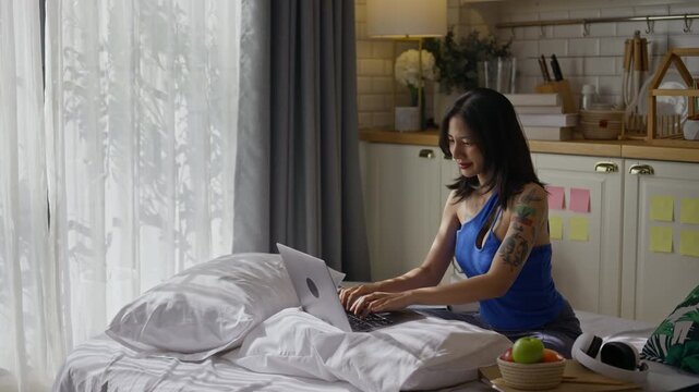 Freelancer woman Gen Z sitting on bed and typing on a laptop in apartment.asian female remote work in kitchen at home.digital nomad,online work,remote working and video conference training concept