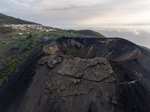 Aerial view of the volcanic crater on La Palma with rugged black lava formations, surrounding green vegetation, and a small town in the distance under a cloudy sky in the Canary Islands, Spain.