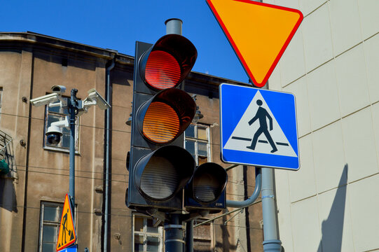 Urban city street intersection featuring an illuminated red traffic light, blue pedestrian crossing sign, and yield warning sign against a bright clear sky