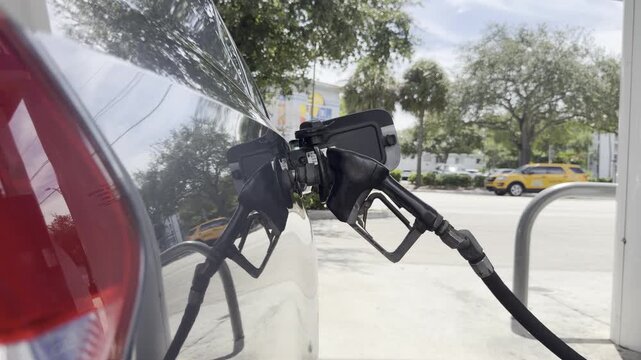 Static 4K extreme close-up of a gas pump nozzle inserted into a car's tank, actively pumping fuel with no hands in frame. Cars pass behind. Shot on a tripod, raw unedited video.