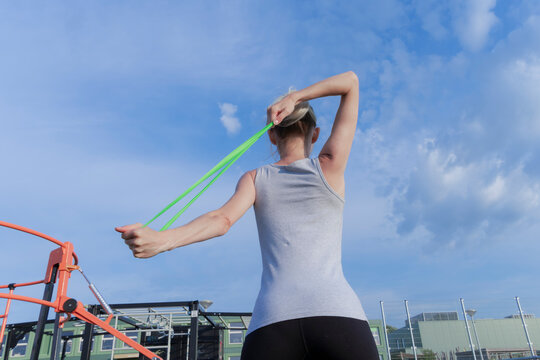 Blonde woman stretching green resistance band overhead against blue sky back view