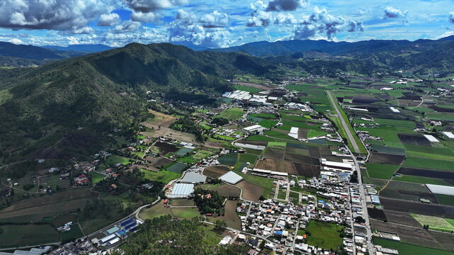 Aerial view of the Portezuelo valley featuring agricultural fields, greenhouses, and a small town nestled among mountains under a blue sky in Constanza, La Vega, Dominican Republic.