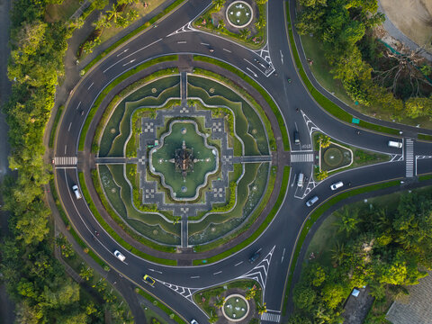 Aerial view of a circular roundabout with a central monument mandala and fountain surrounded by lush greenery and roads in Kecamatan Kuta Selatan, Bali, Indonesia.