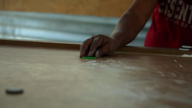 Young man in red essentials shirt flicking carrom striker with focused expression inside warm lit village room near corrugated tin wall