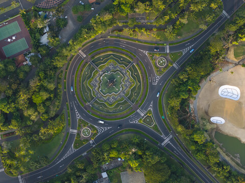 Aerial view of a circular roundabout with lush green landscaping and geometric paths surrounded by trees and a golf course in Kecamatan Kuta Selatan, Bali, Indonesia.