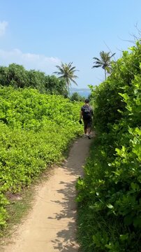 Man with backpack walking along narrow path surrounded by tropical greenery toward ocean