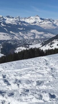 Vertical 9:16 video showing a panoramic winter landscape from a snowy summit near Megeve in the French Alps. The scene begins with a close-up of textured, trampled snow.