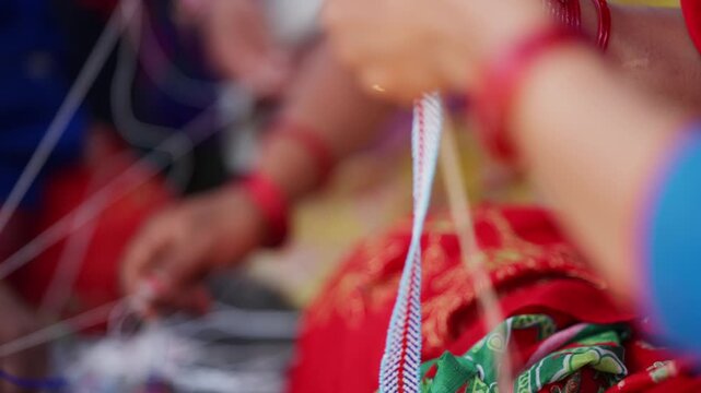 Skilled hands adorned with red bangles braiding pote bead thread strands in extreme close up with softly blurred festival background