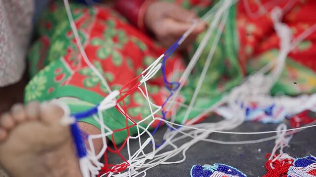 Weathered hands with red bangles threading blue pote bead string over vivid red and green floral fabric beside blurred bare foot