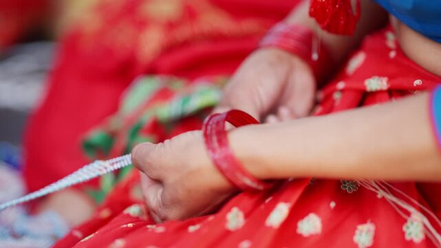 Close up of hands with red bangles and dangling red tassel earring threading pote bead string against vibrant red sari background