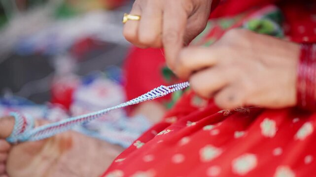 Hands with layered red bangles carefully guiding pote bead string while seated in vivid red traditional sari during far west Nepali craft work