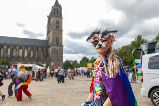 Masked therian girl in colorful rainbow cape poses during CSD celebration in Magdeburg, Germany, with cathedral behind. Scene reflects youth subculture, fandom, identity, pride, creativity, community