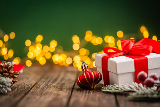 A Christmas background featuring a white gift box tied with red ribbon beside a pinecone, red ornaments, and frosted berries on rustic wood, lit by warm twinkling lights above with copy space.