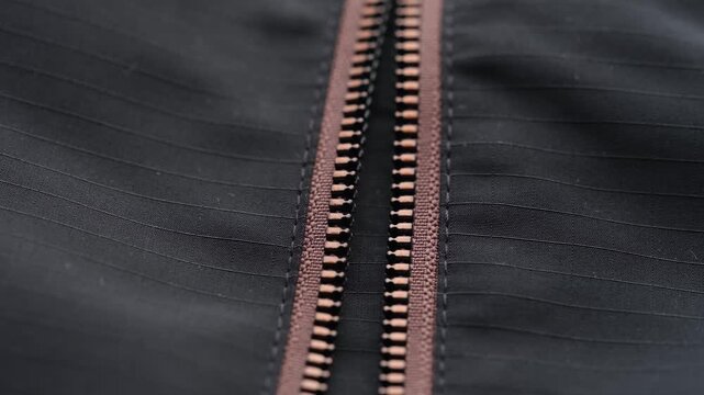 A macro moving video of an open zipper on a black waterproof ripstop jacket with brown tape details.