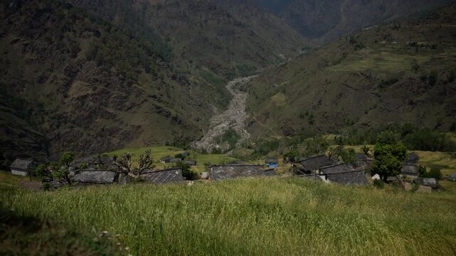 Traditional stone village with dark slate rooftops sits below a dramatic mountain gorge with a dry rocky riverbed winding through steep forested slopes