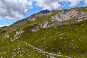 Fototapeta premium Scenic hiking trail on green grassy slope with rocky mountains under cloudy blue sky, trekking to Lancebranlette peak, Little St Bernard Pass, Aosta Valley, Italy