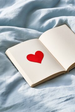 Open cream notebook with a red heart-shaped paper cutout on a blank page, resting on softly rumpled pale blue bedding in gentle morning light