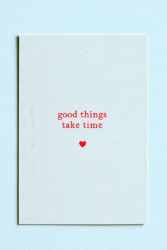 Minimalist motivational poster featuring the phrase good things take time in red serif font on a clean powder-blue background with a small red heart below
