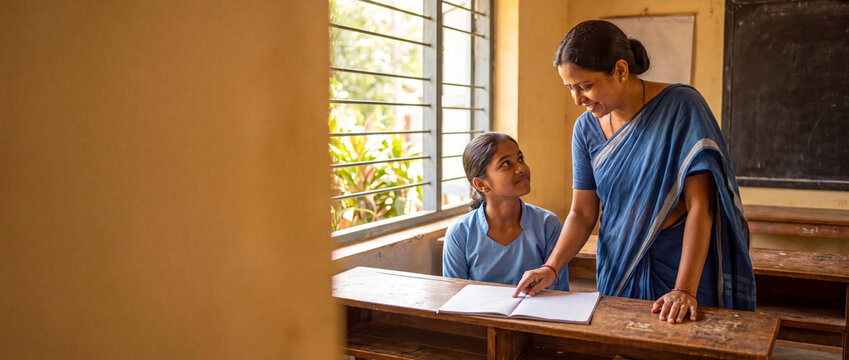 South Asian female teacher in a saree helping a young girl read at a wooden desk in a rural classroom with copy space.