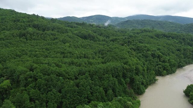 Aerial 4K footage over Tekhuri river near Martvili, Georgia: drone climbs toward a forested hill along the riverbank with rolling mountains behind. Overcast morning after rain.