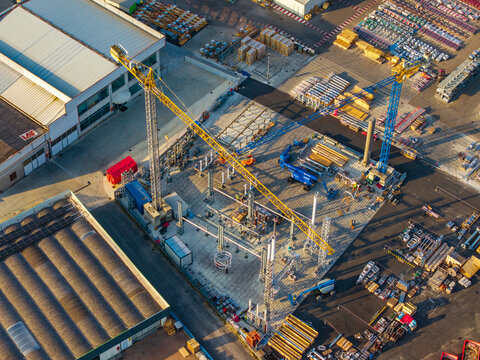 Aerial view of a construction site featuring yellow and blue tower cranes, industrial warehouses, and stored materials in Noventa di Piave, Veneto, Italy.