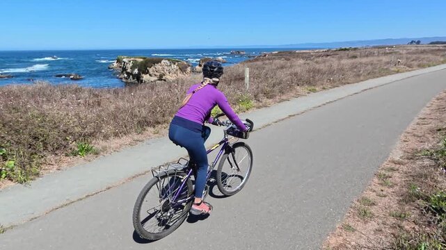 Mature woman enjoys biking on a paved trail next to the ocean in Fort Bragg, California on a nice day.
