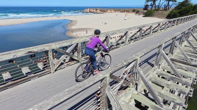A senior woman rides her bike on a wooden bridge next to the ocean in Fort Bragg, California near Mendocino. The weather is clear and sunny.