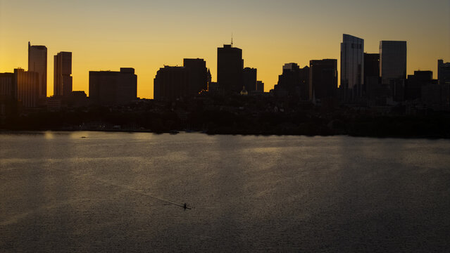 Aerial view of the Boston skyline at sunset with a lone rower on the Charles River, featuring silhouetted skyscrapers and calm water in Boston, Massachusetts, United States.