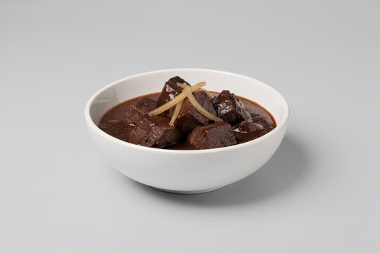 Vlaamse Stoofkarbonaden in white bowl, rich dark brown sauce, chunks of beef, glossy sheen, pop of color from lemon peel. Concept of beef beer stew with onions mustard.
