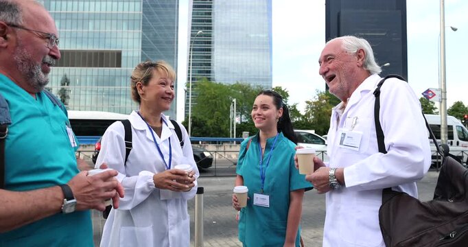 Professional group of smiling doctors and nurses in uniform talking and relaxing together while having a coffee break outdoors, with a contemporary clinic building in the background