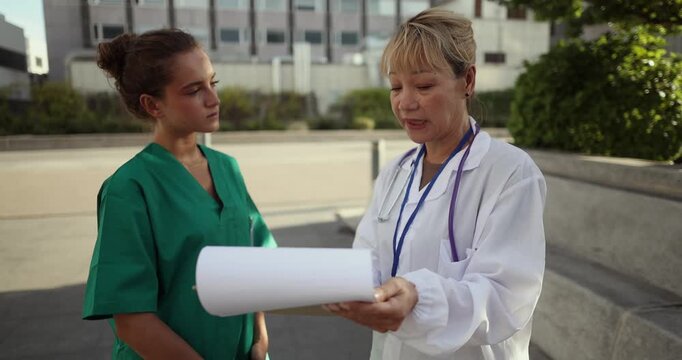 Experienced female doctor with a stethoscope providing guidance and instruction to a younger nurse intern, both discussing a patient's medical chart on a clipboard outside the hospital building