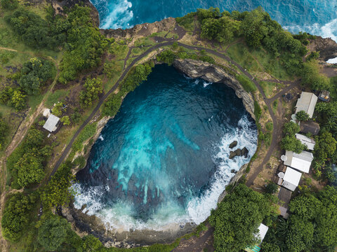Aerial view of Broken Beach featuring a natural rock archway over turquoise sea water, surrounded by lush green cliffs and walking paths in Nusa Penida, Bali, Indonesia.
