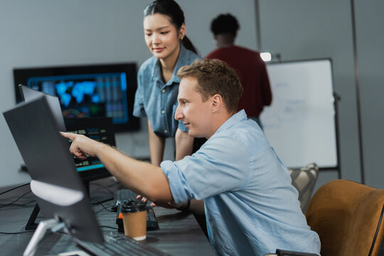 Focused developer types code on a dual monitor workstation while a colleague stands behind him, working through a software build in a dark modern technology office.