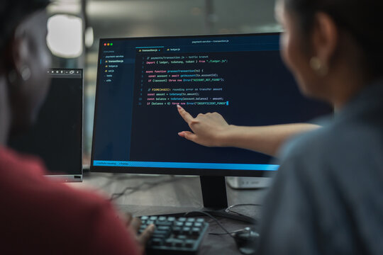 Male and female developers discuss programming solution together, pointing at lines of code on monitor while working as small software team in dim office