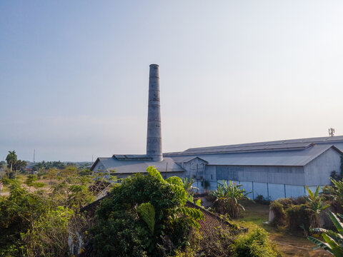 Aerial view of an old industrial factory with a tall brick chimney, surrounded by lush tropical vegetation and trees in the rural landscape of Banyumas Regency, Central Java, Indonesia.
