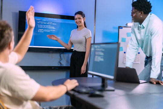 Team member raises hand with question while woman explains code on wall screen, software developers in training session in blue lit modern office with whiteboard behind