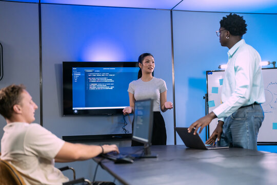 Three colleagues review software project in conference room, woman presents code on wall screen while man works at laptop and seated developer listens at long table