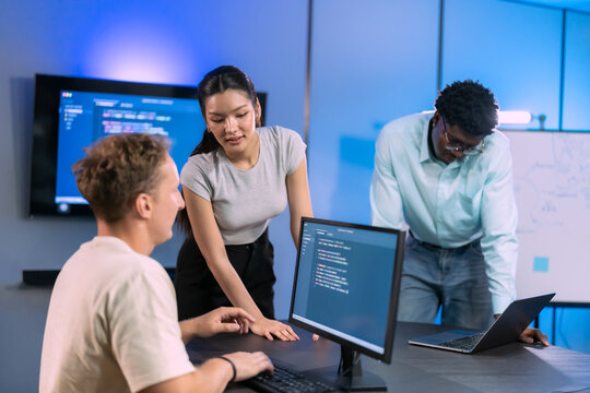 Three diverse software developers smile and talk together around their desks, with code on the wall screen and an agile flow diagram on the whiteboard behind them.